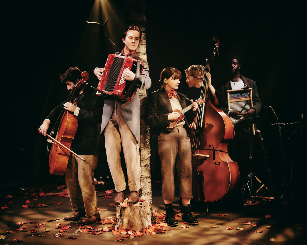 Five musicians on stage with red accordion, double basses, and other instruments against dark background with warm stage lighting.