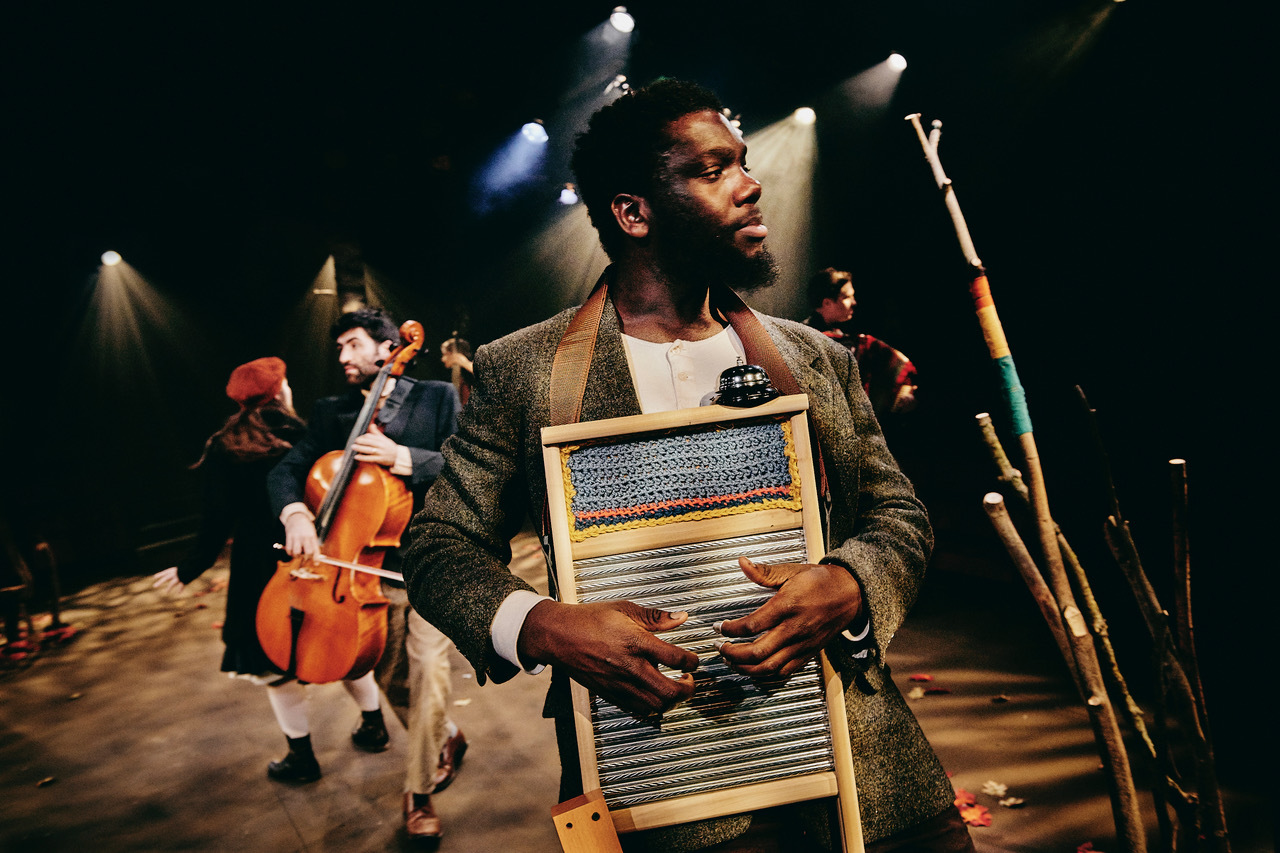 Black musician holding washboard instrument on stage with dramatic lighting, other performers with string instruments in background.