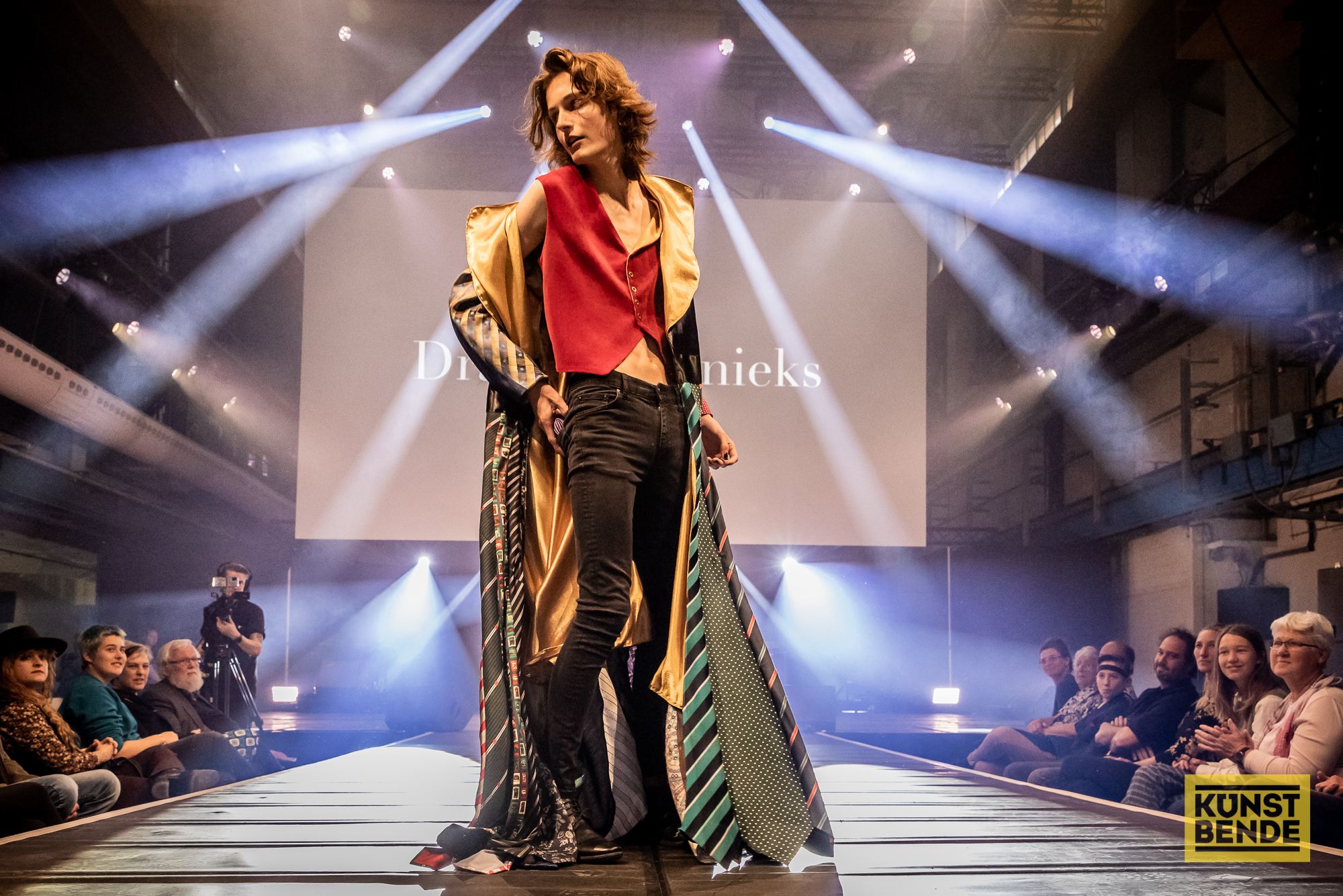 A model posing at the end of a catwalk with spotlights shining all around. People are sitting either side of the runway.