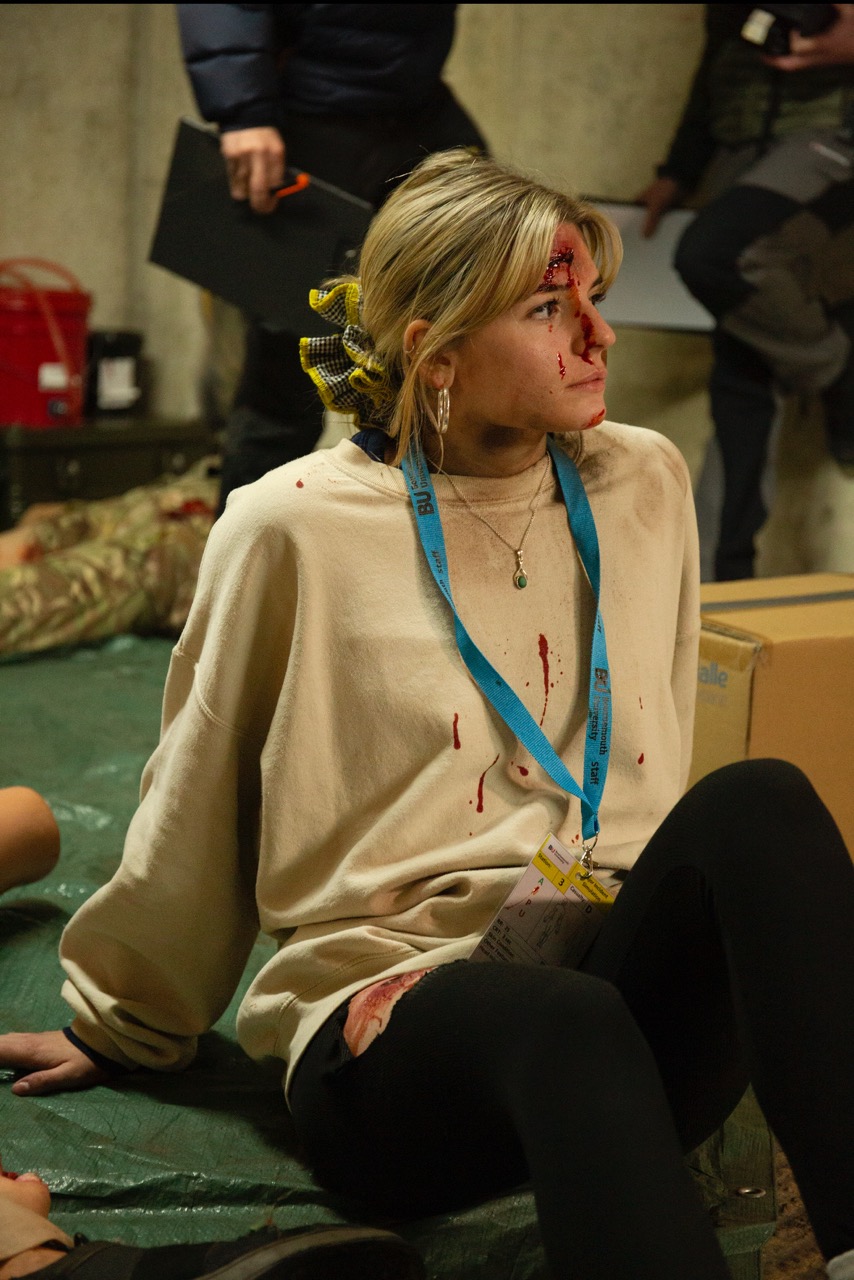 A woman wearing a beige sweater sits on the floor with visible blood on her clothing and face. She has a yellow scrunchie in her hair, and a blue lanyard around her neck. The background shows other individuals and supplies in a dimly lit room.