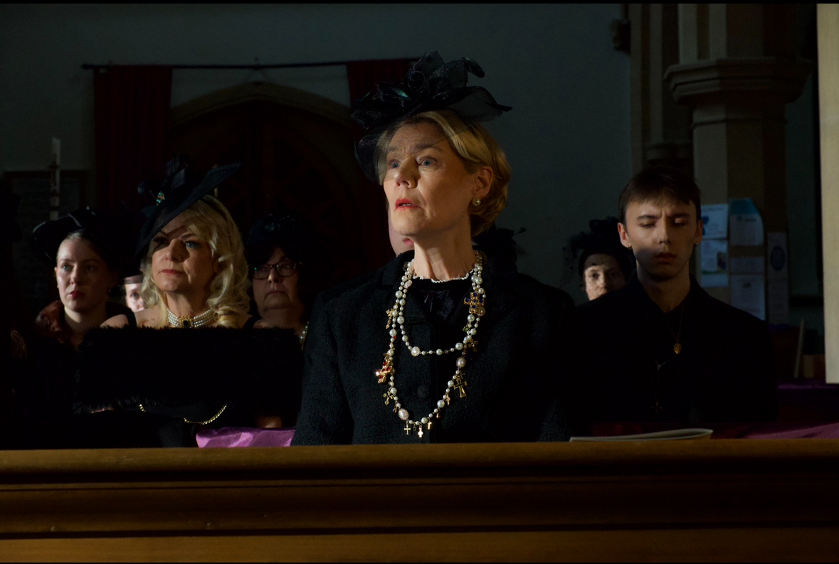 A group of people dressed in black attire sitting in a church during a service. The focus is on a woman in the foreground wearing a black outfit and ornate necklace, with several others wearing hats in the background.