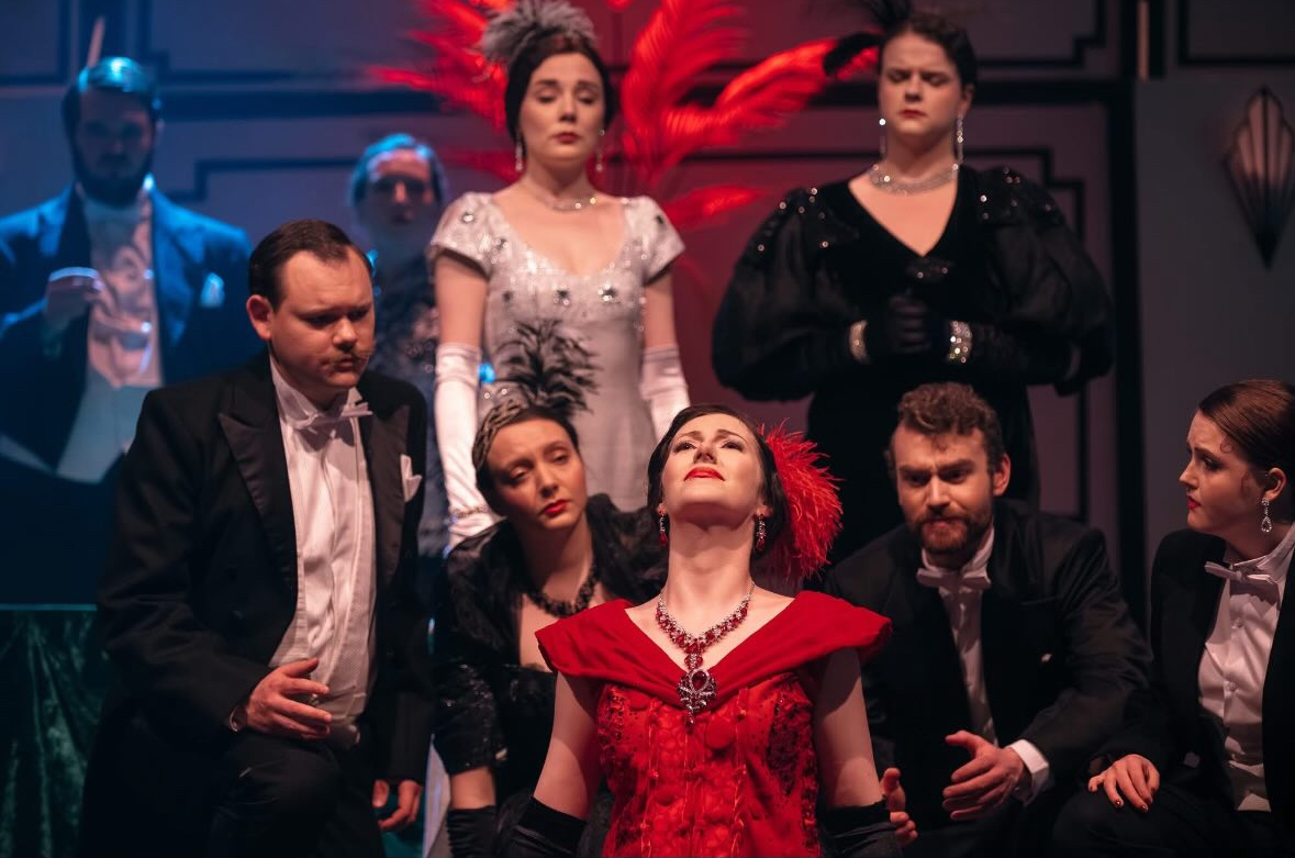 A theatrical scene featuring several performers in formal attire, including a woman in a red dress with a feathered headpiece at the front. The background features dramatic lighting and ornate decorations, enhancing the performance's atmosphere.