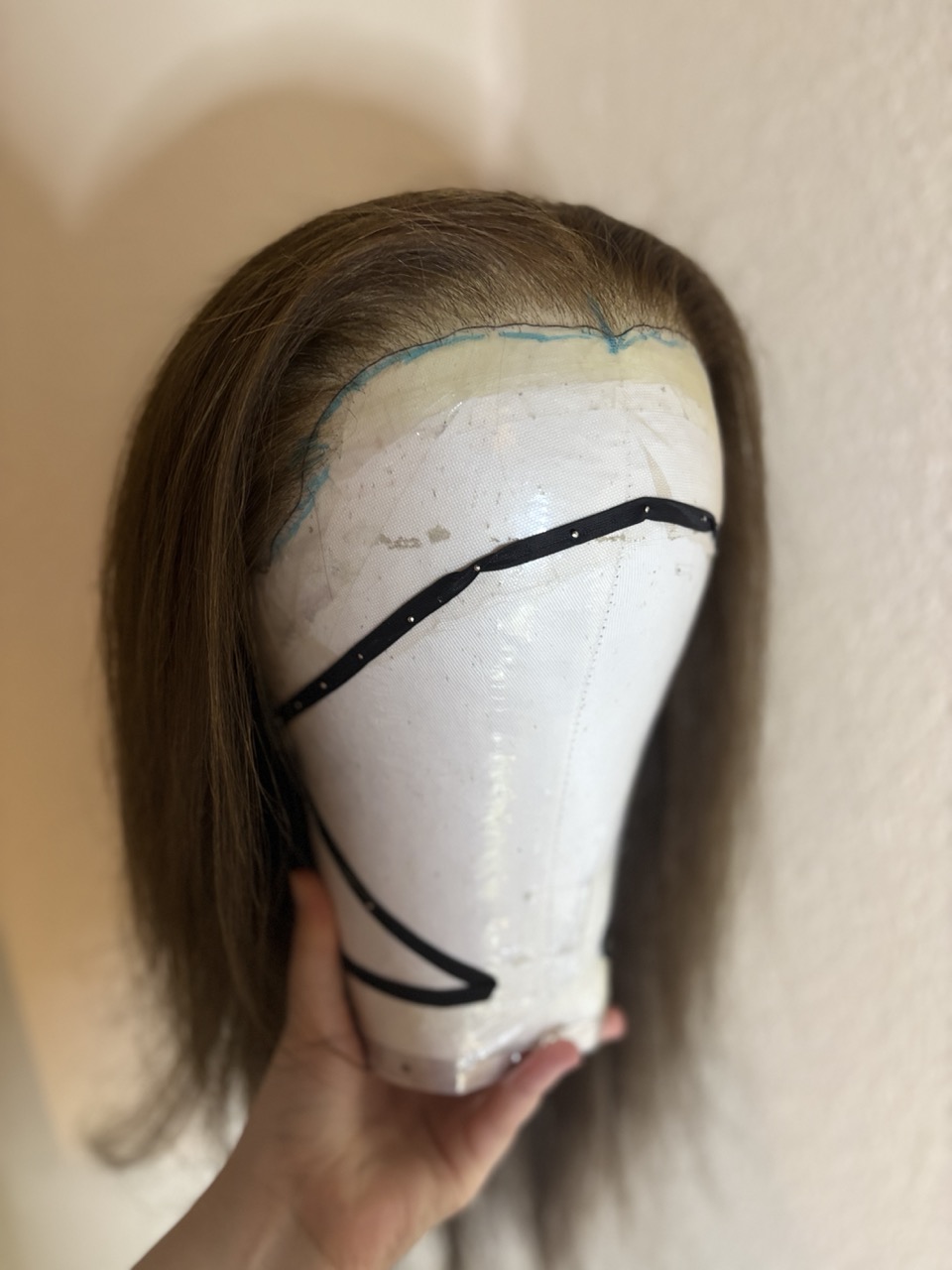 A clean, white wig head with light brown hair attached, showing a smooth surface with contour markings. The wig head is held in a hand, with a plain wall serving as the background.