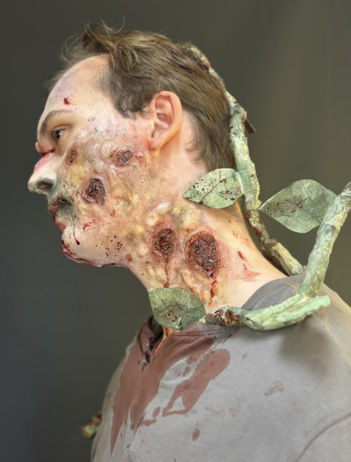 A side profile of a man with a severely distressed appearance, featuring textured skin lesions and a discoloured complexion. Green vines with leaves wrap around his neck, contrasting the grey shirt he is wearing.