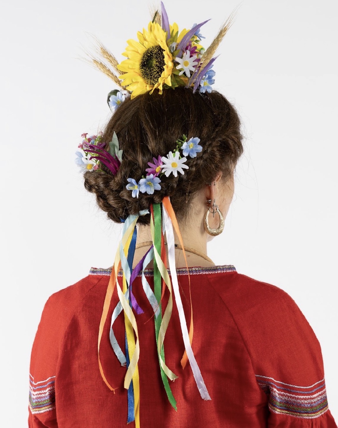 A woman wearing a traditional red embroidered garment with a decorative floral headdress. The hair is styled in a braided bun adorned with colourful ribbons and a mix of sunflowers, daisies, and other flowers.