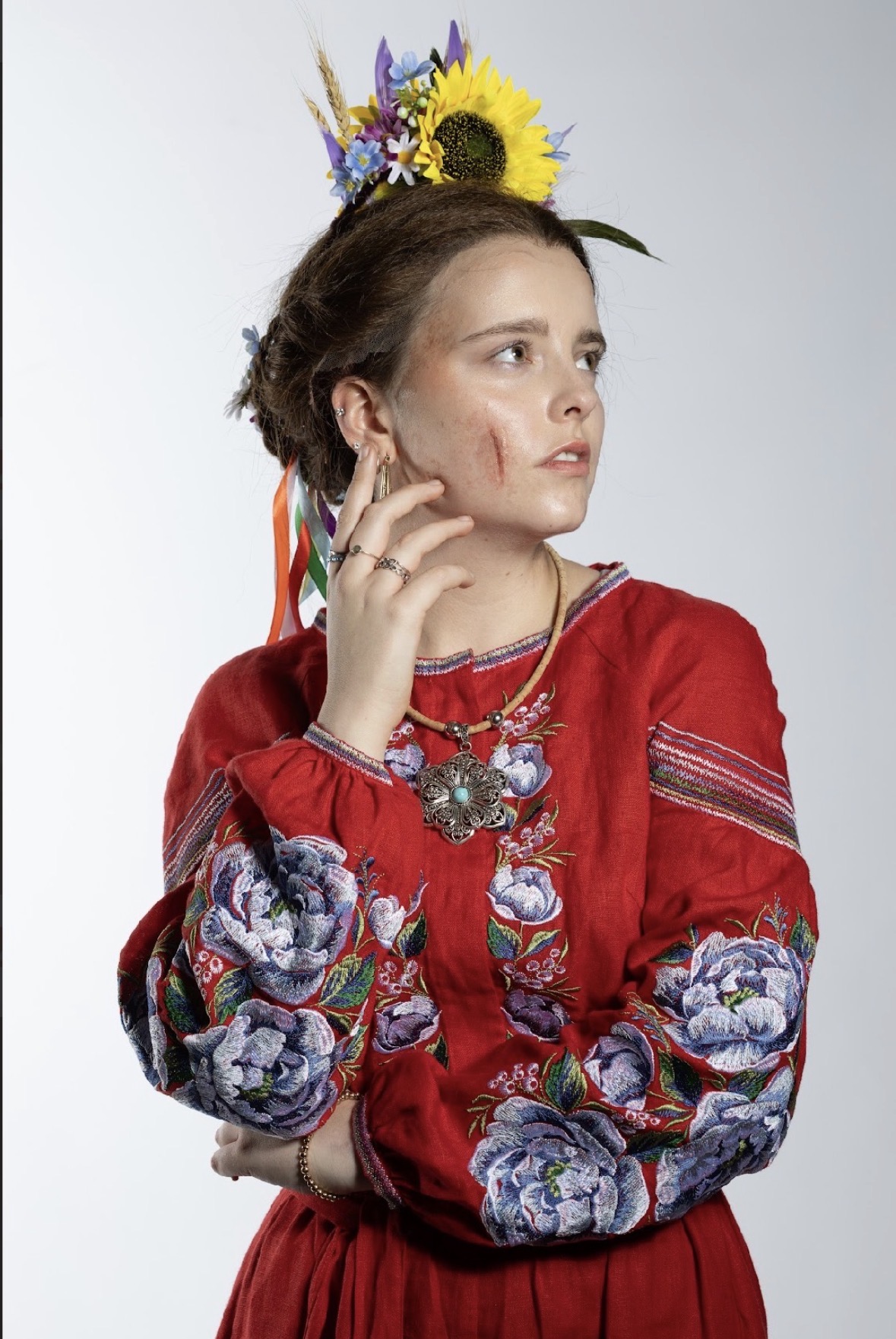 A woman wearing a red traditional dress adorned with floral patterns stands with her hand raised near her face. She has a floral crown on her head and holds a necklace, with a subtle scar visible on her cheek.