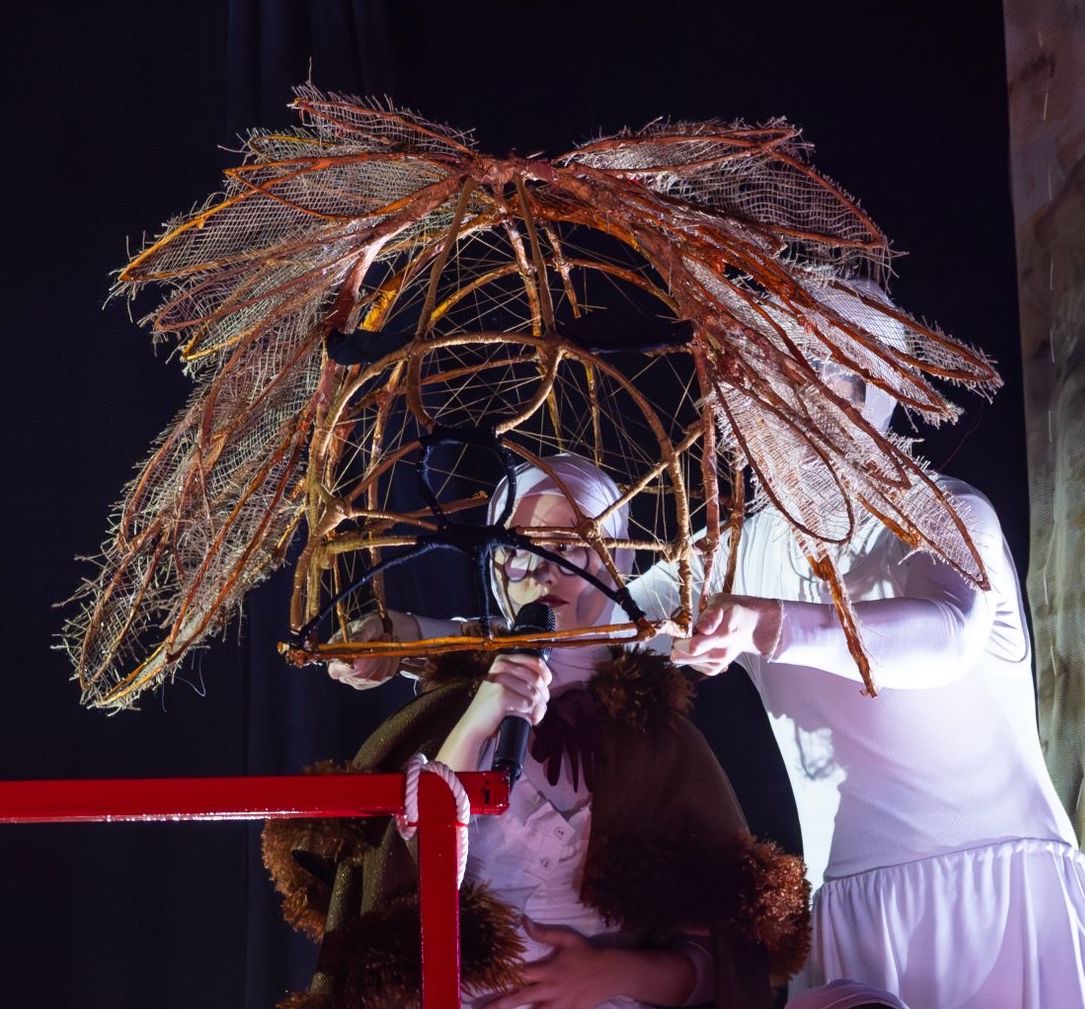 A performer wearing a brown cloak sits on a platform while another performer in white holds a large, elaborate headpiece made of twigs and netting above them. The background is dark, with hints of light highlighting the figures.