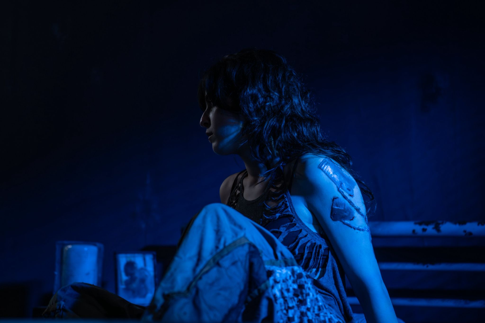 A woman with long, dark hair sits on a step, partially illuminated in blue light. She wears a sleeveless top, revealing a tattoo on her upper arm, while the background features indistinct shapes and shadows.