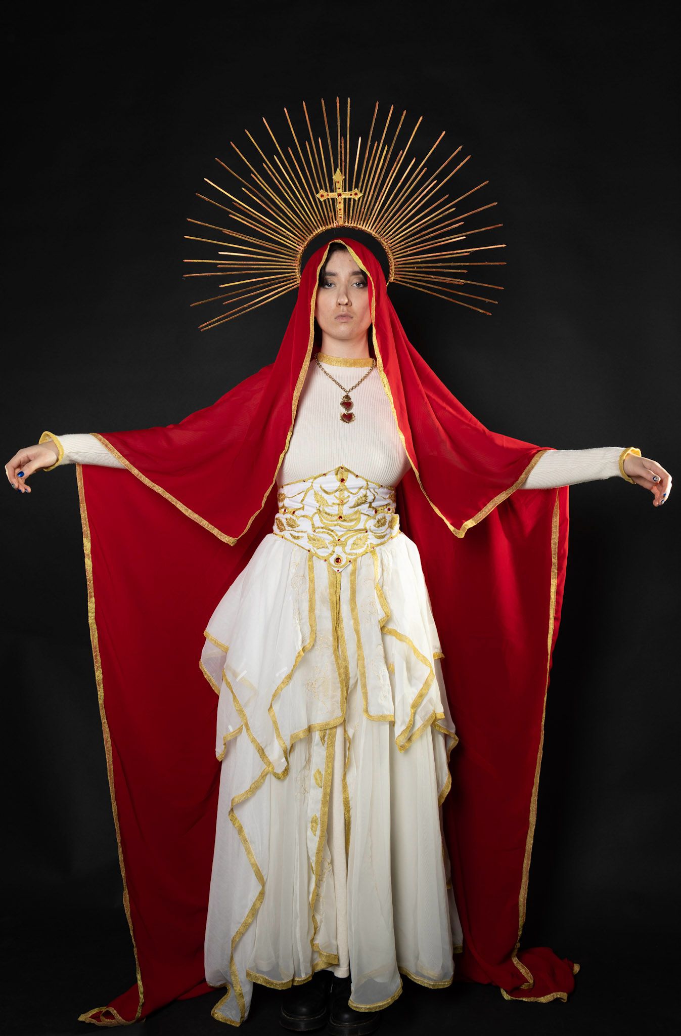 A woman dressed in a white gown with gold trim and a flowing red cape stands against a black background. The outfit features a decorative gold belt and a headdress of golden rays, adding a regal appearance.