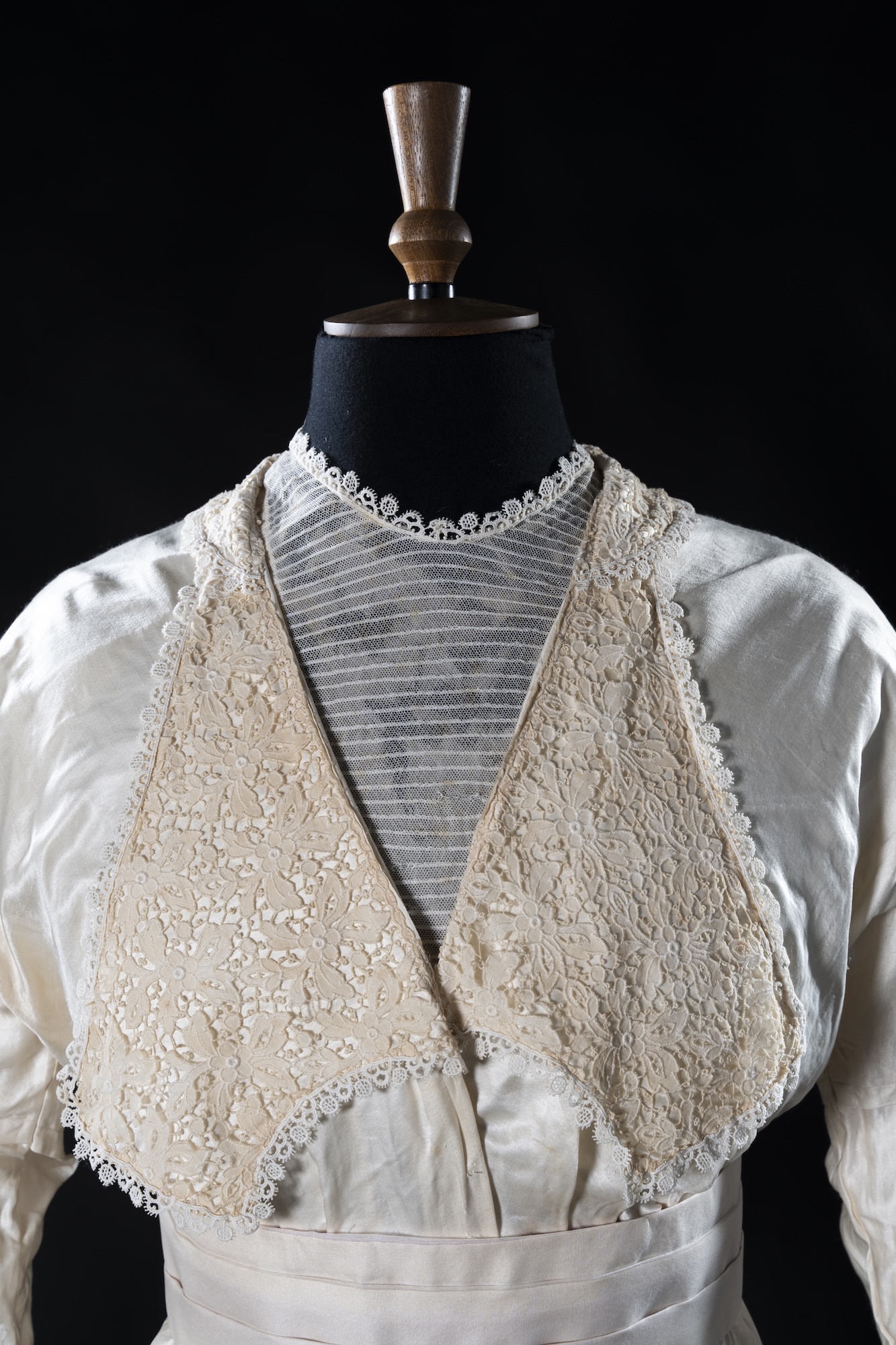 A vintage white satin dress with lace panels and a striped lace bodice is displayed on a mannequin against a black background.