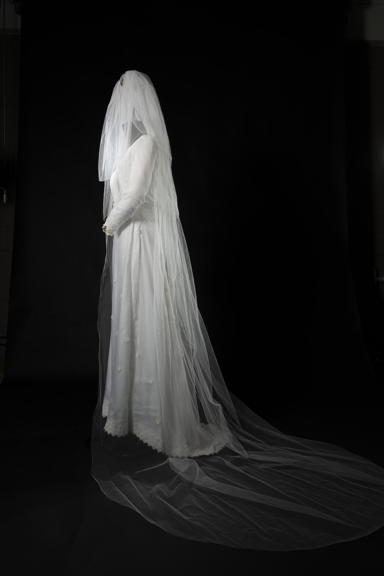 A woman in a long white wedding dress and veil stands in profile against a black background, with a flowing train trailing behind her.