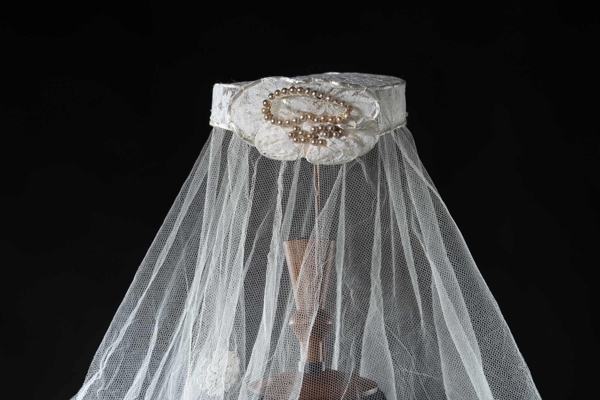 A white lace bridal veil with a beaded floral detail drapes over a mannequin-like form against a black background.