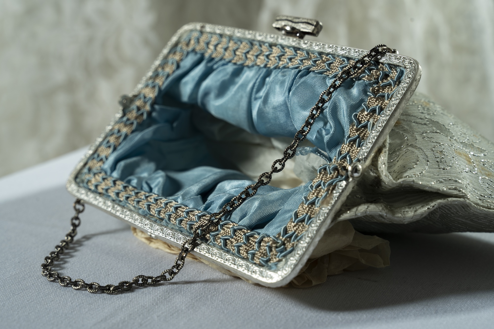 A silver clutch handbag with a black chain strap and blue satin lining rests on a grey surface, with folded fabric behind it.