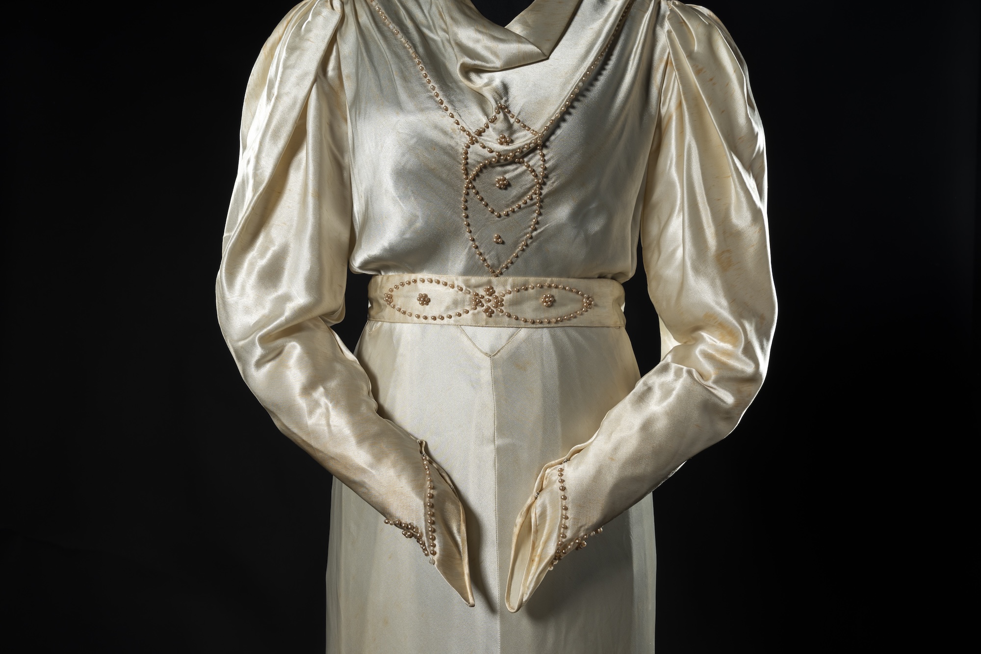 A cream satin dress with long puffed sleeves and beaded heart details, shown against a black background.