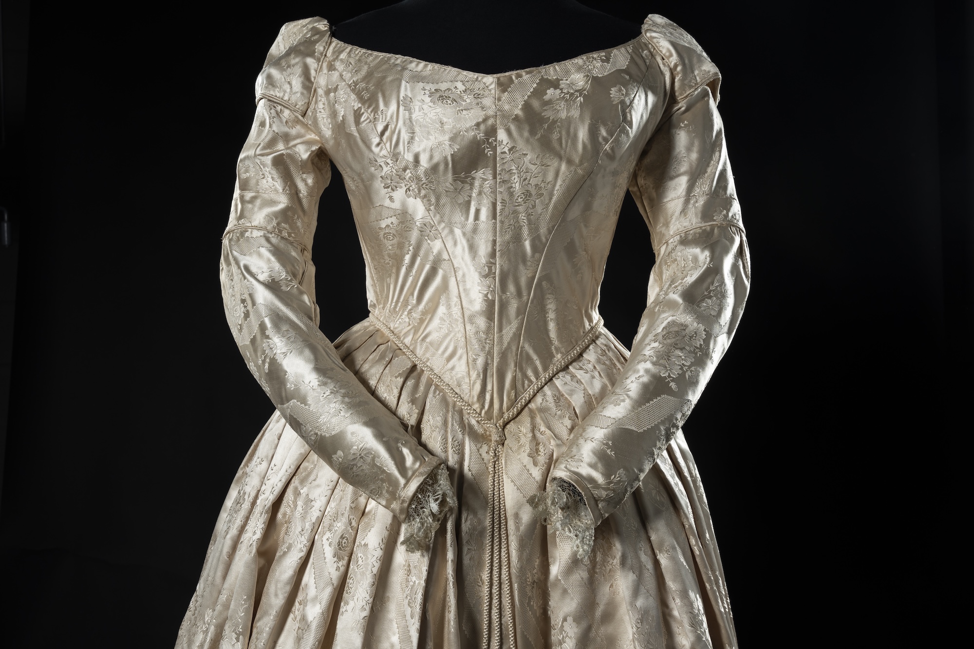 A cream satin long-sleeved dress with a fitted bodice and full pleated skirt, displayed against a black background.