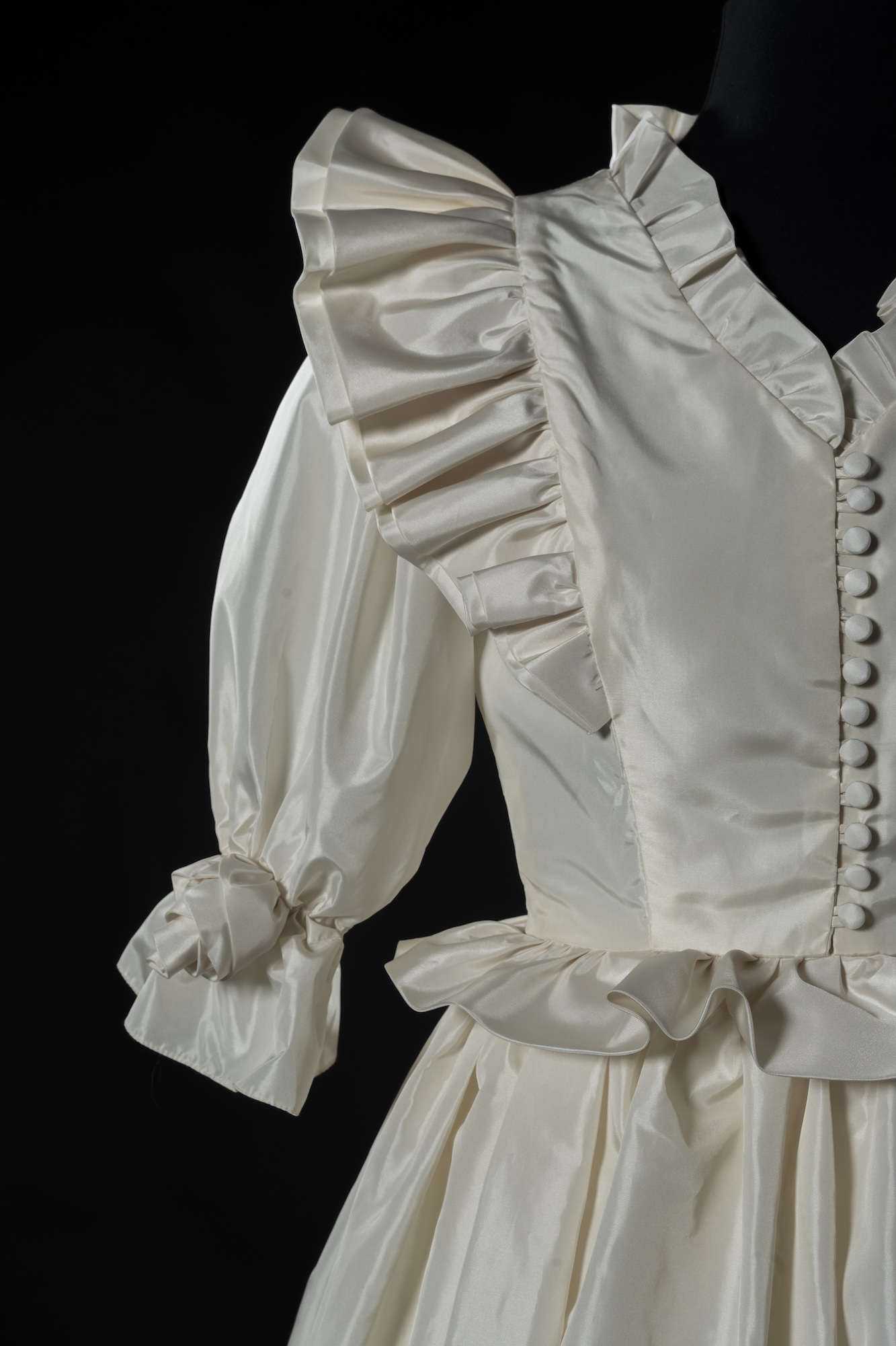 A cream Victorian-style dress with ruffled trim, puffed sleeves and button detailing, shown against a black background.