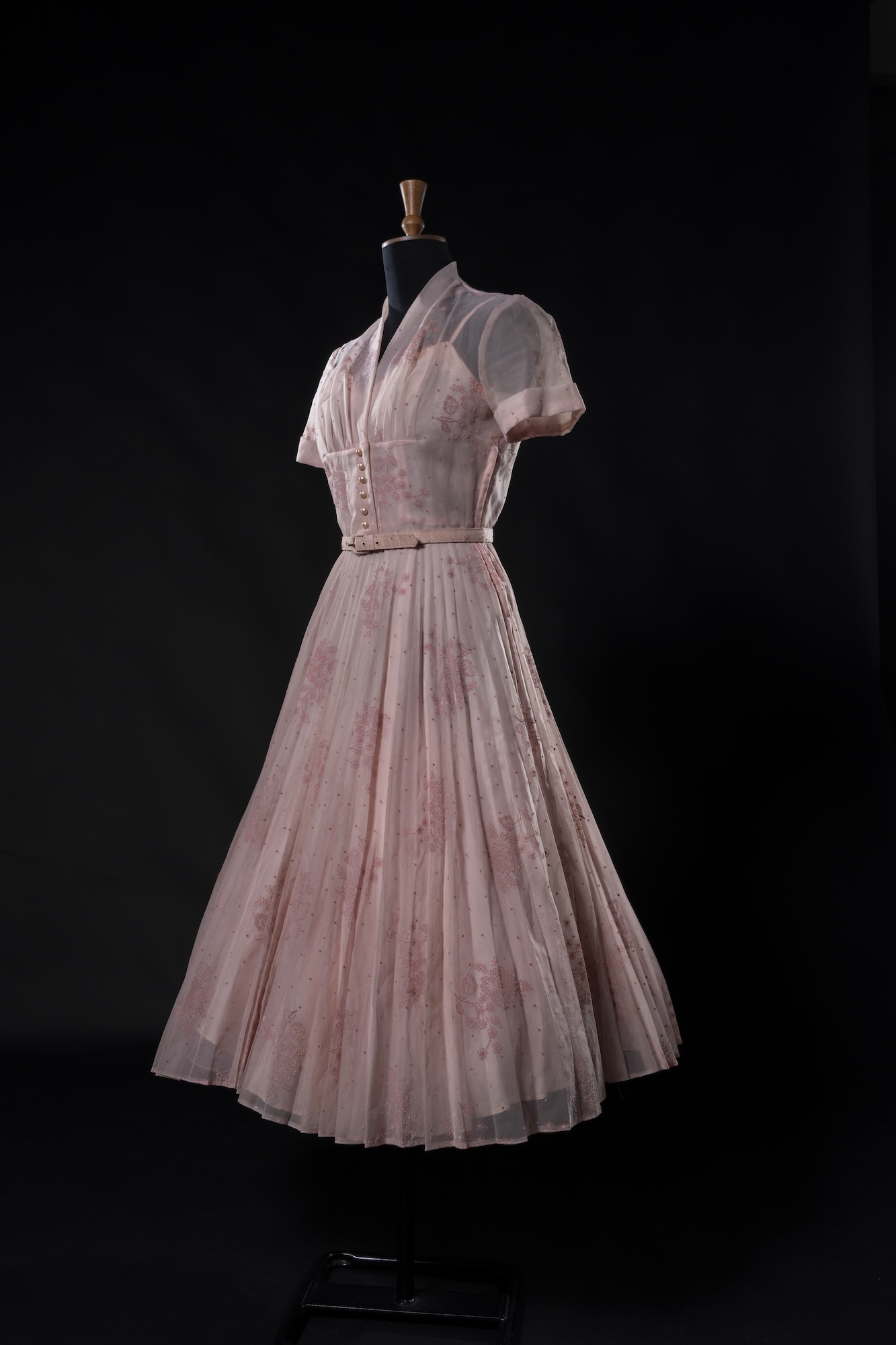 A pale pink pleated dress with short puff sleeves is displayed on a mannequin against a black background.