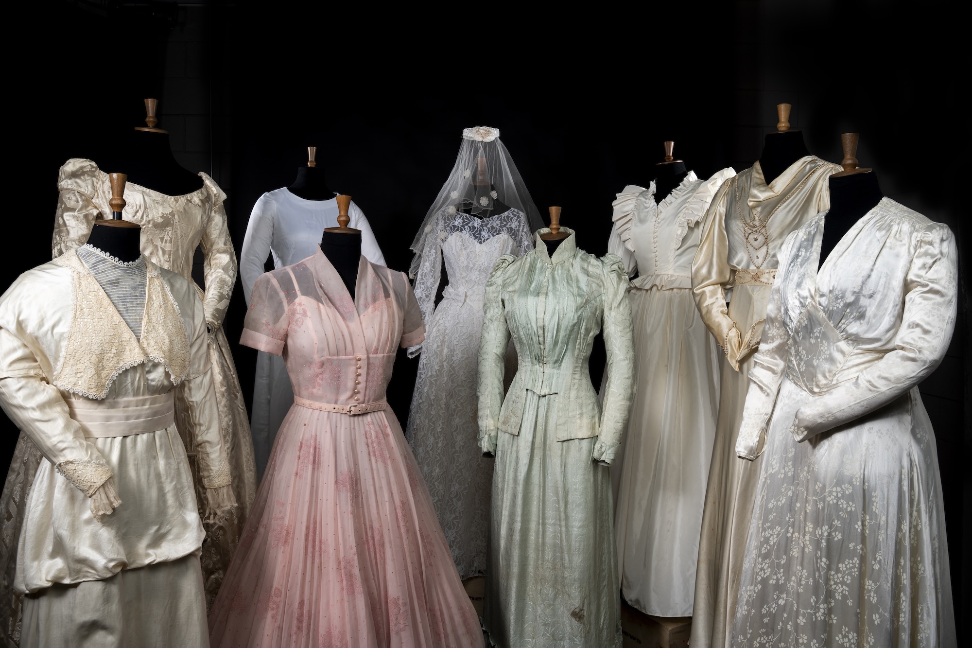 A row of vintage wedding dresses on dress forms, including white, pink and pale green gowns against a black background.