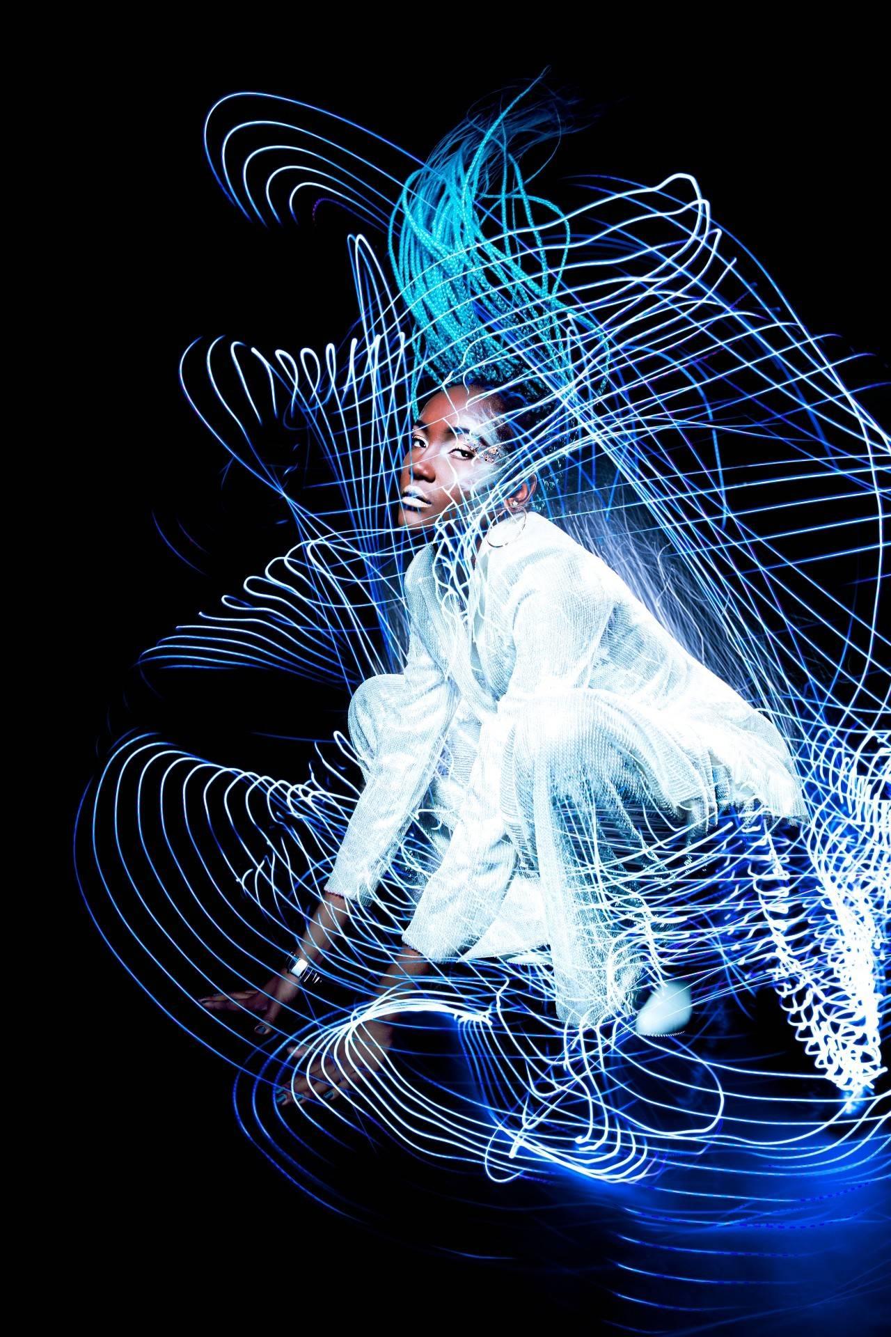 A model in white costume crouches down with luminescent lines all around them.