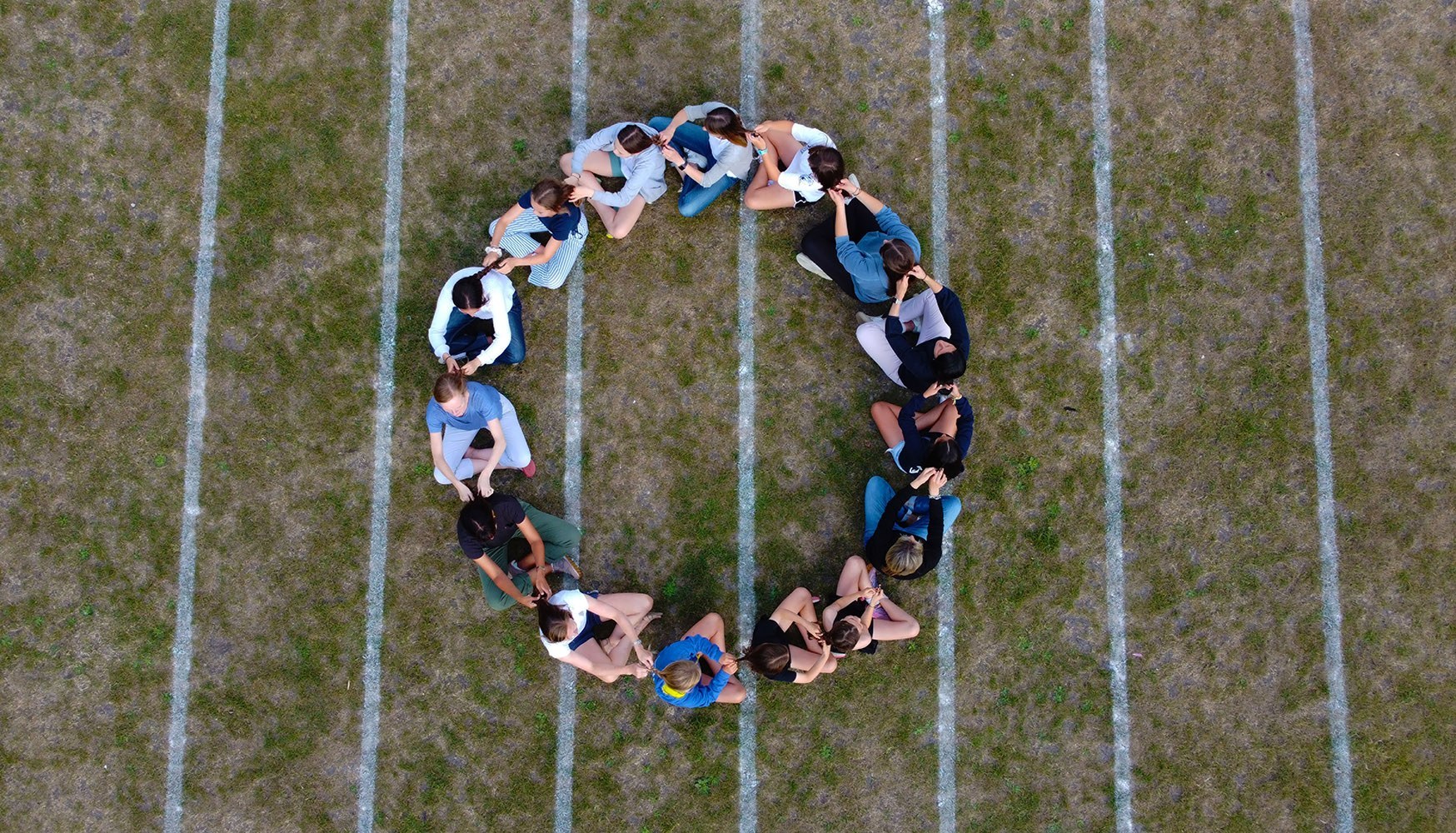 Drone photographic image taken from above of women and children sat cross legged in a circle plaiting each other's hair to create a continuous loop of plaiting.  They are sat on a school field with white stripes on it.