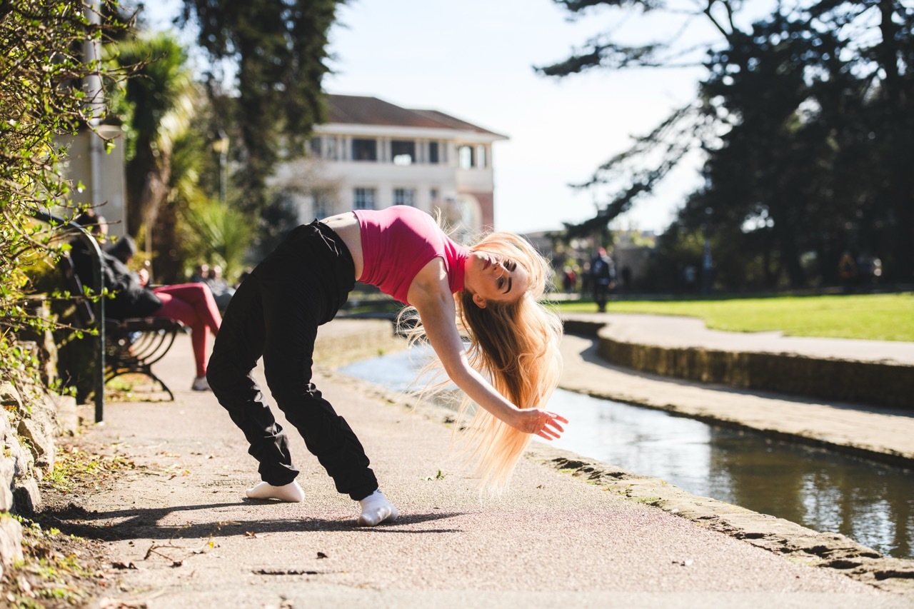 Woman in red top bending over concrete edge of water feature, long blonde hair hanging down, reaching towards water in park setting.