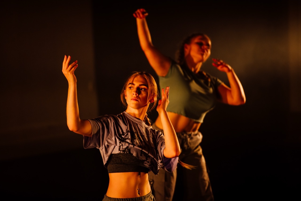 Two performers on stage with orange stage lighting. Blonde woman in grey crop top with raised arms, brunette woman behind with microphone.