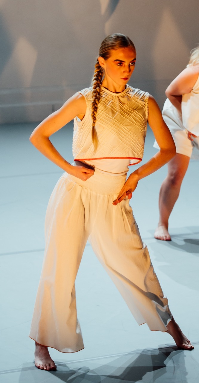 Dancer in white crop top and wide-leg trousers with long plait, hands on hips, orange stage lighting, other performers visible behind.
