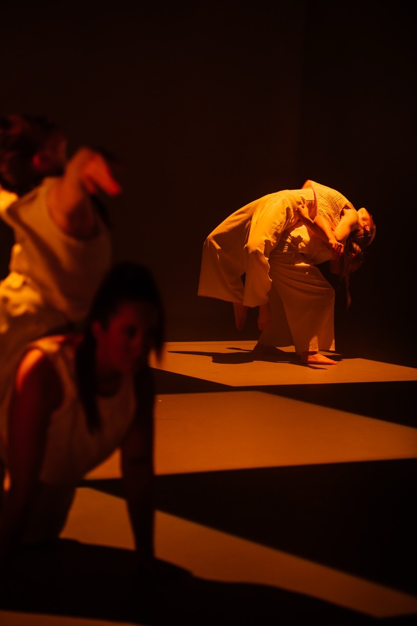 Two figures in white clothing on orange-lit stage, one crouched on platform, another blurred in motion, dark background with warm lighting.