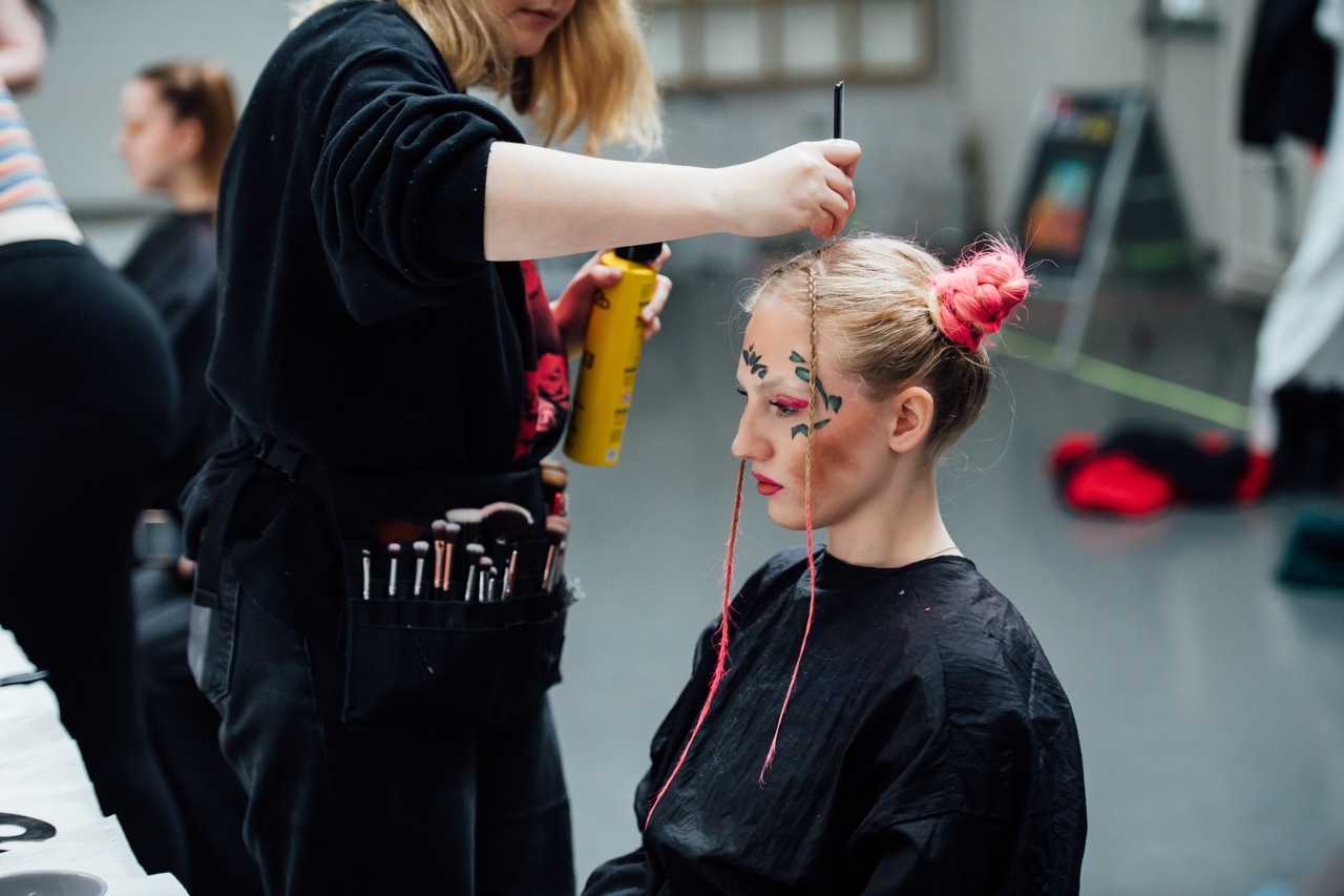 Make-up artist styling blonde hair, using brush on woman wearing black gown with red hair accessories.