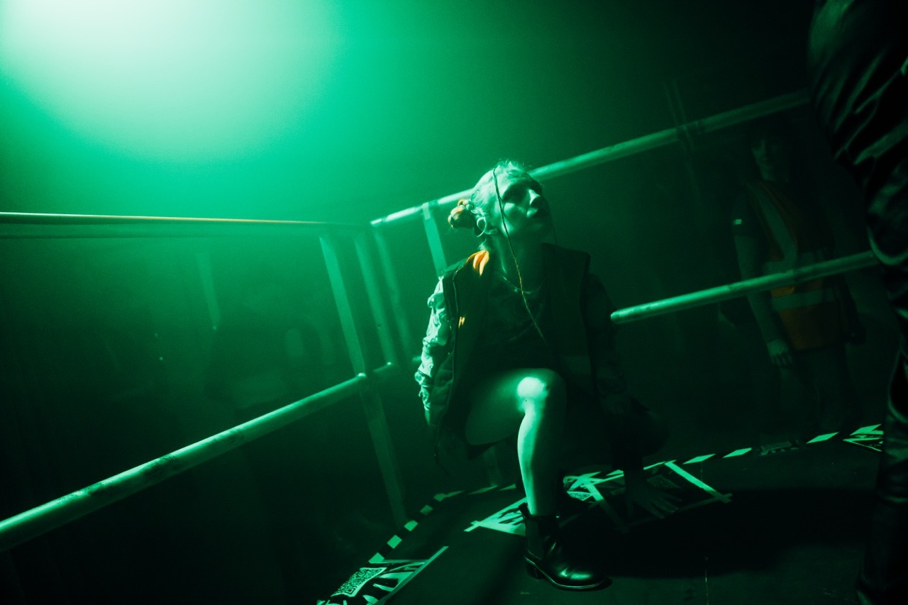 Five dancers on stage jumping upwards with arms extended under blue-green lighting.