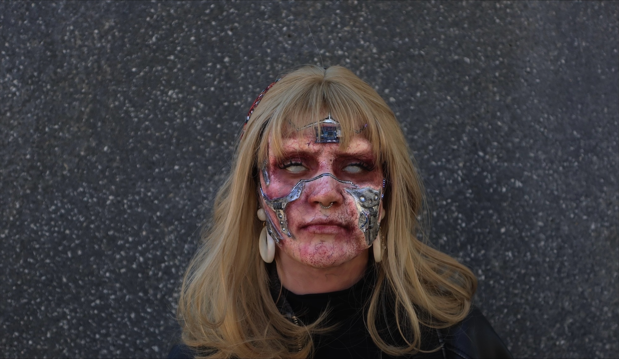 Headshot of a model wearing sci-fi prosthetics; the model is made to look like a corpse with grey eyes and red skin, blonde hair, and metal pieces fused to their cheeks, forehead and across their nose.