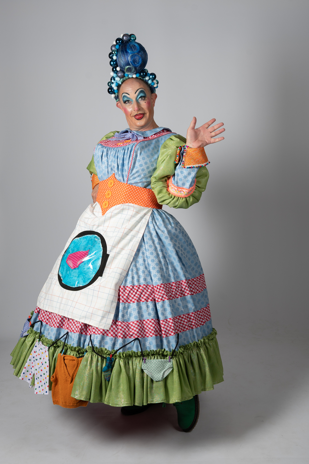 Pantomime dame in theatrical costume with blue hair curlers, colourful patchwork dress, white apron, and exaggerated makeup waving hand.