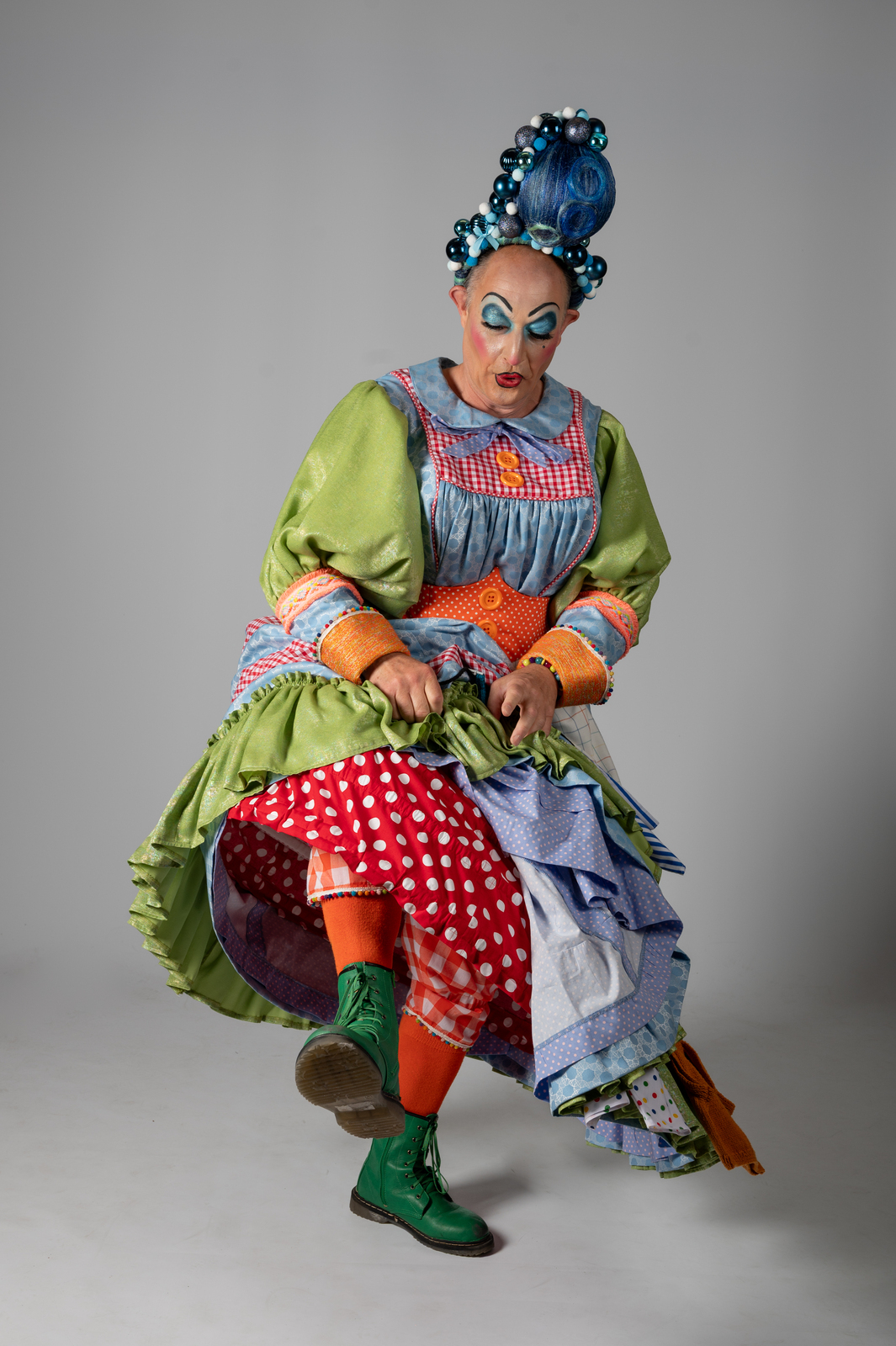 Pantomime dame in colourful layered costume with blue hair, dramatic makeup, green sleeves, checkered apron, polka dot skirt, striped tights.