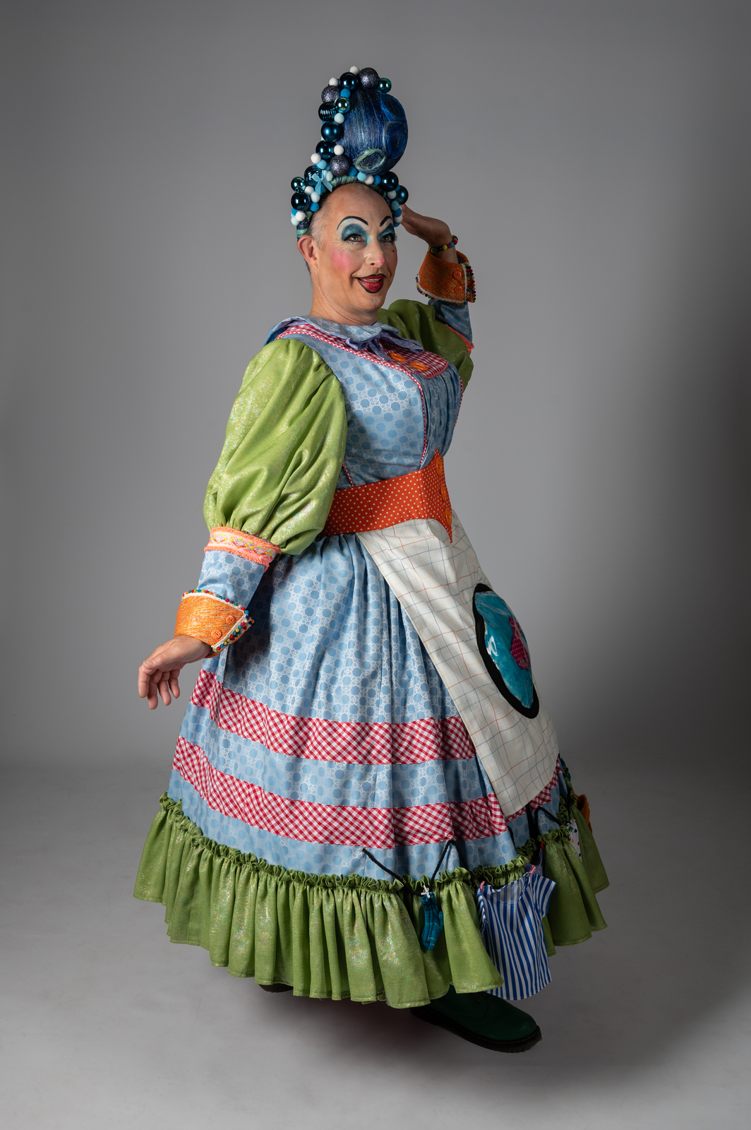 Pantomime dame in colourful costume with blue hair, layered dress in blue, green, red and white patterns, white apron, posed theatrically.