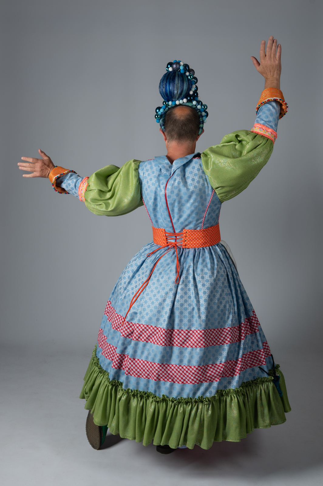 Pantomime dame in traditional costume with blue bodice, green sleeves and skirt, red trim, arms raised, back view against grey background.