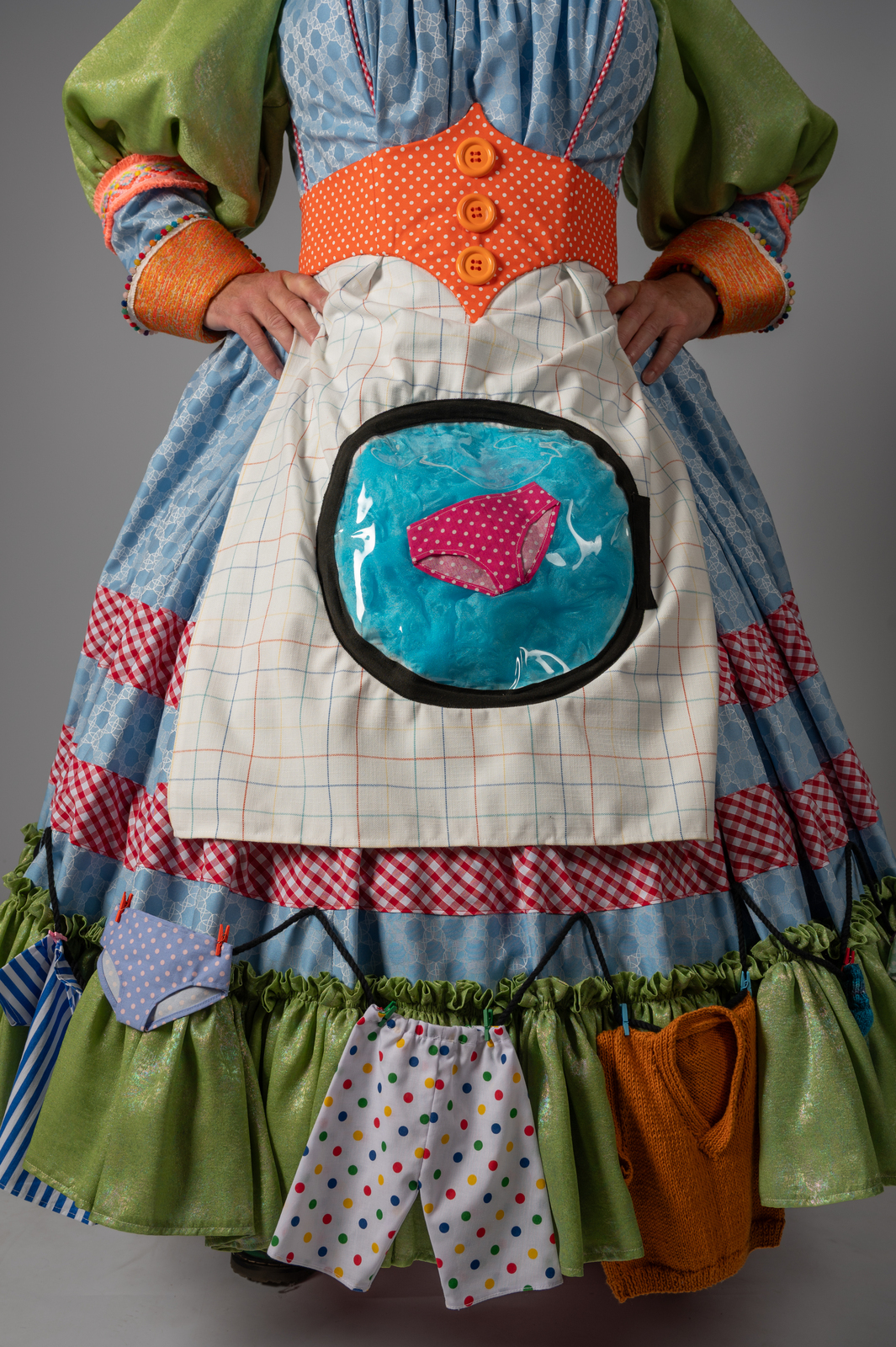Pantomime dame wearing patchwork dress with green sleeves, blue bodice, orange corset, white apron featuring circular window with turquoise and pink fish design.
