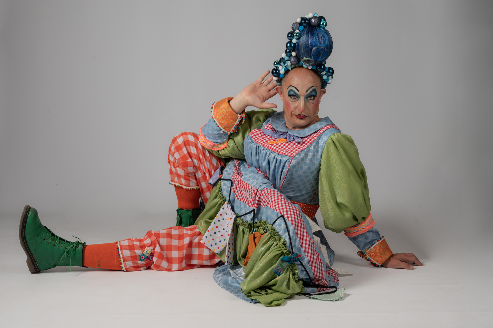 Pantomime dame in colourful patchwork costume with blue hair sits on grey background, wearing green boots and red gingham trousers.