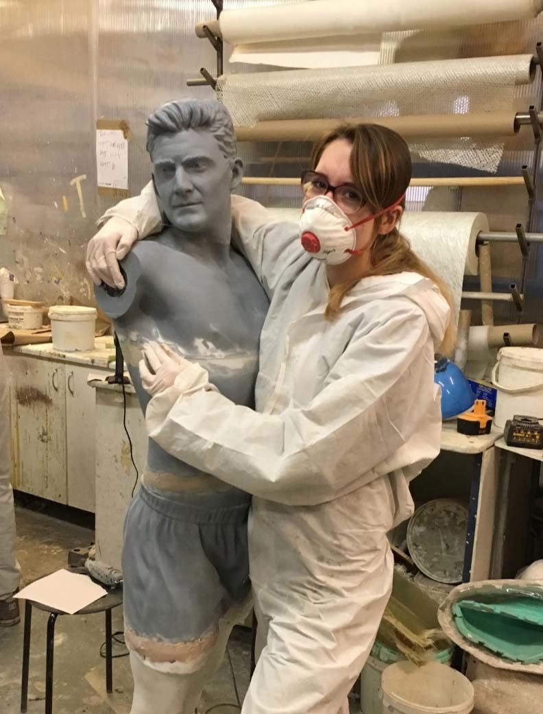 A girl in a white paper coverall, stood next to a statue sculpture she is working on