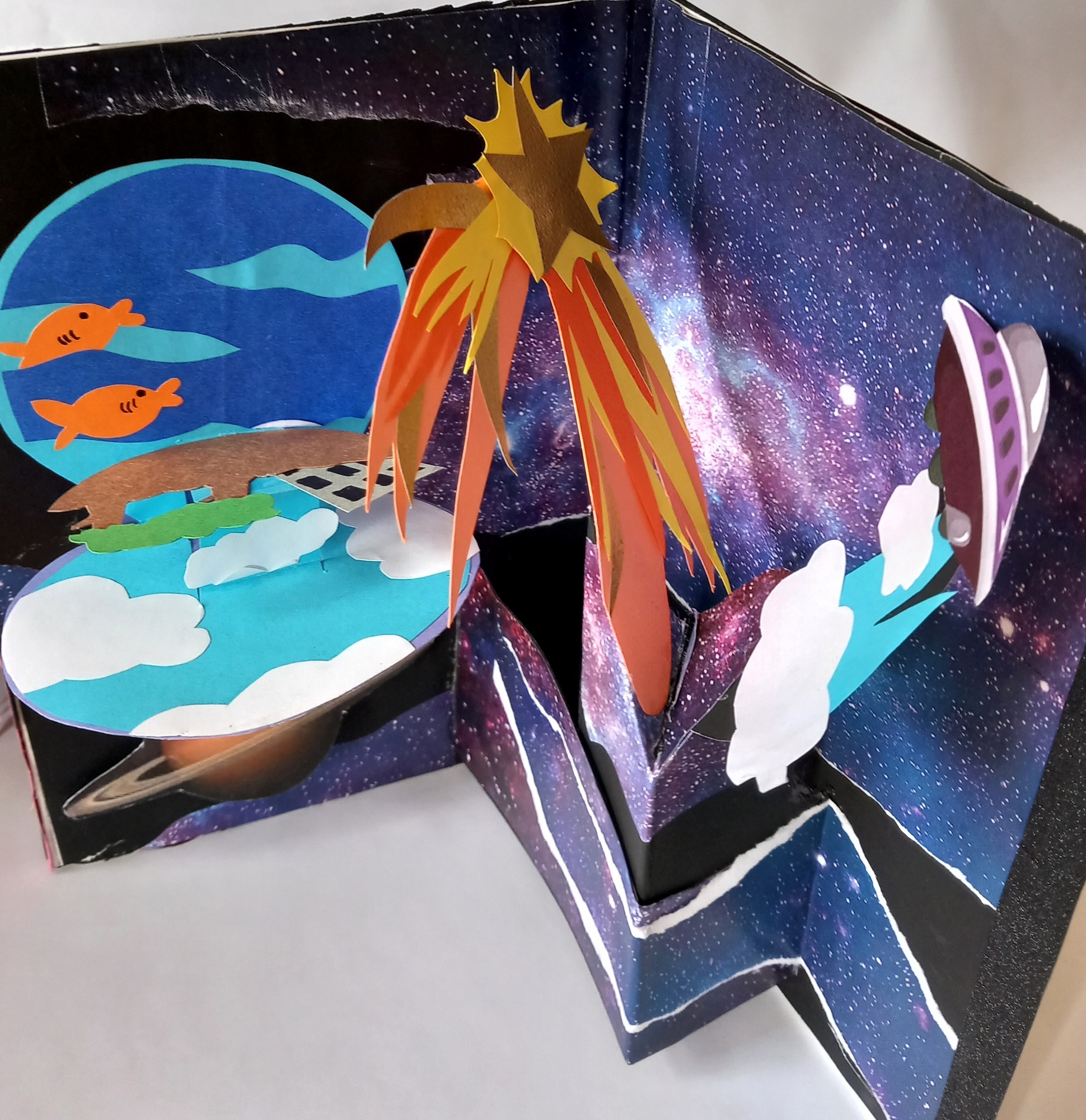 Paper craft diorama with dark blue starry background, yellow-orange star shape, blue circle with white clouds, and white rabbit silhouettes