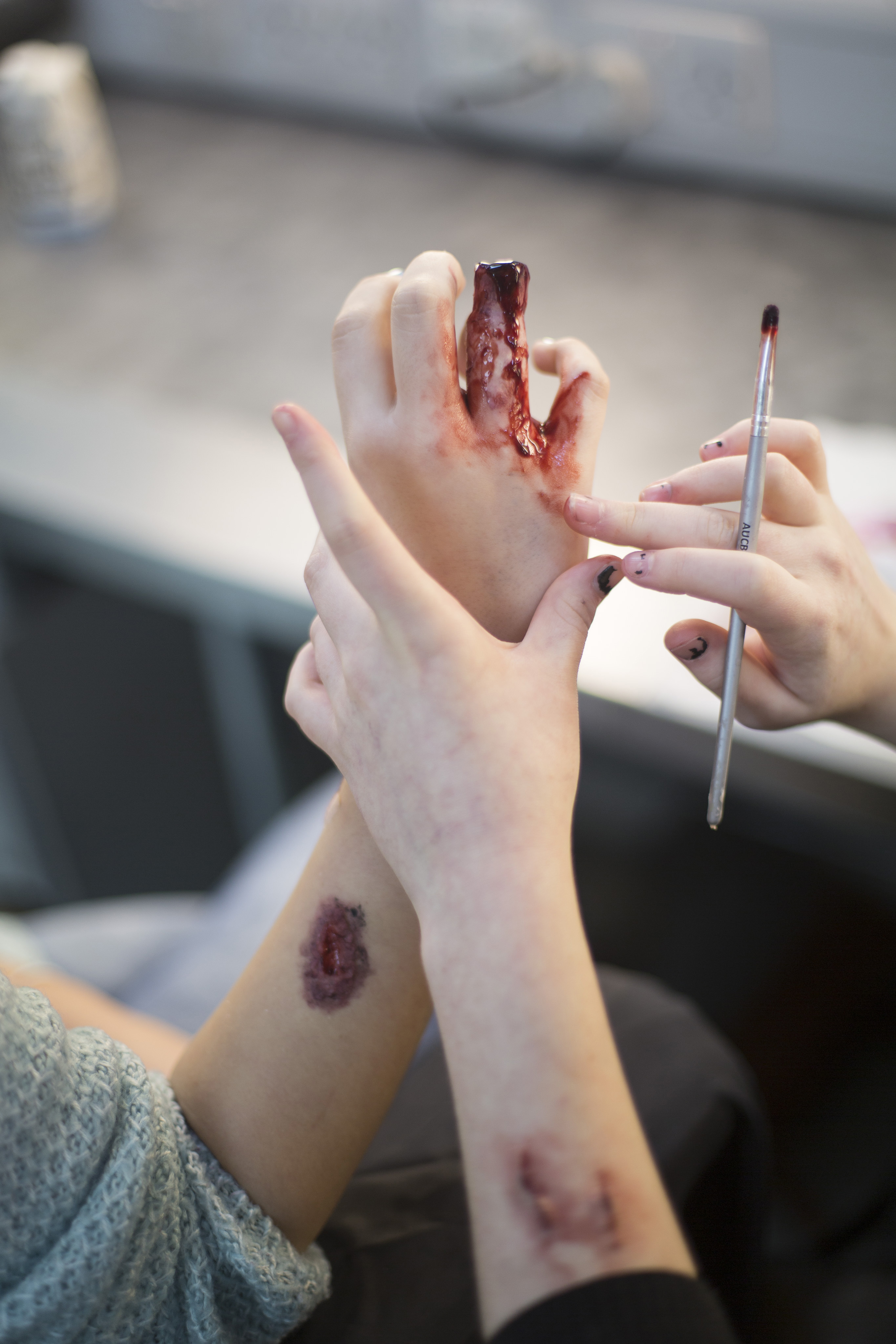 Hands applying dark red theatrical makeup or special effects blood with a brush, showing simulated wounds on palm and forearm.