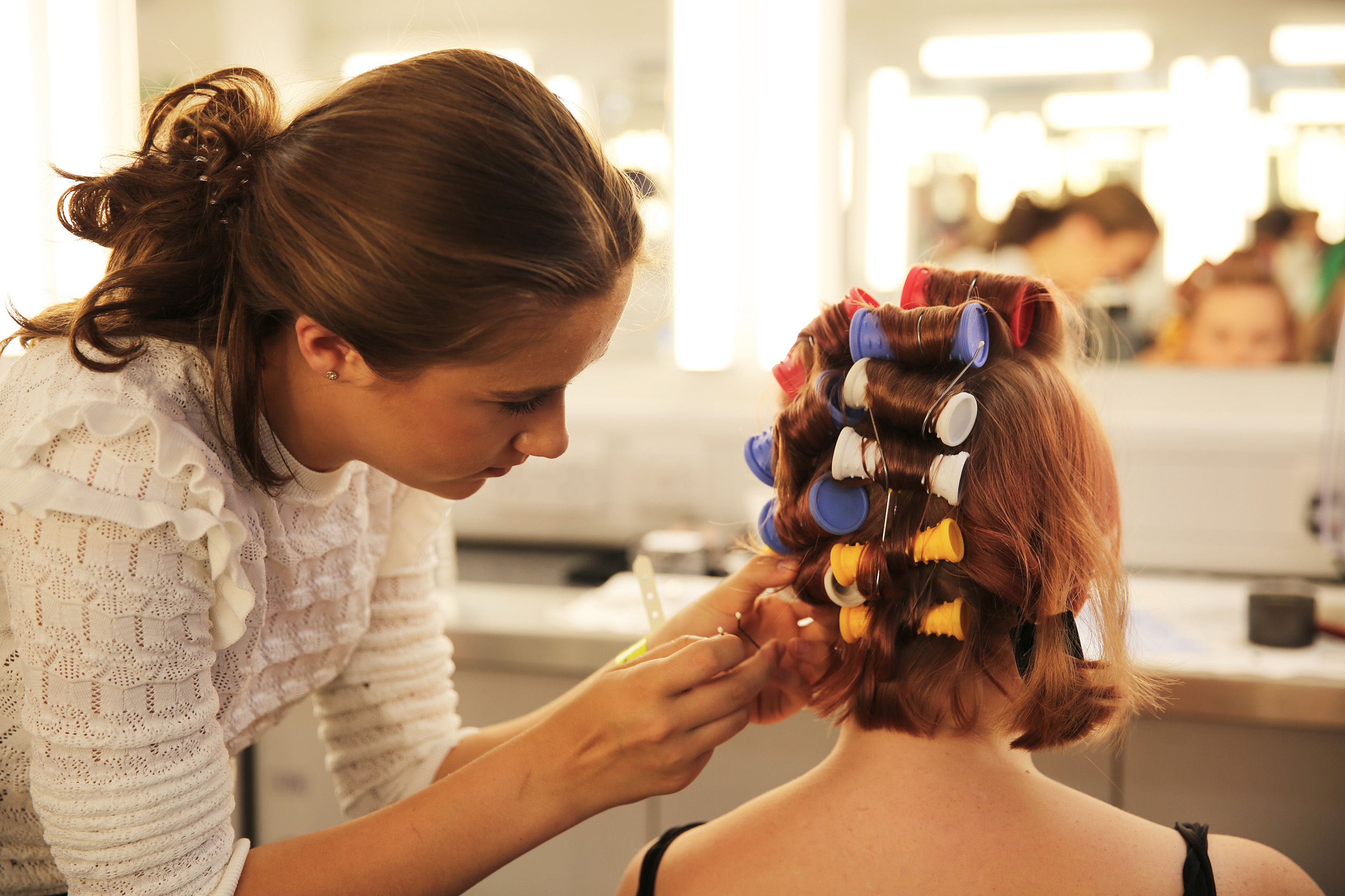 A student is styling another student's hair. They have hair curlers in their hair.