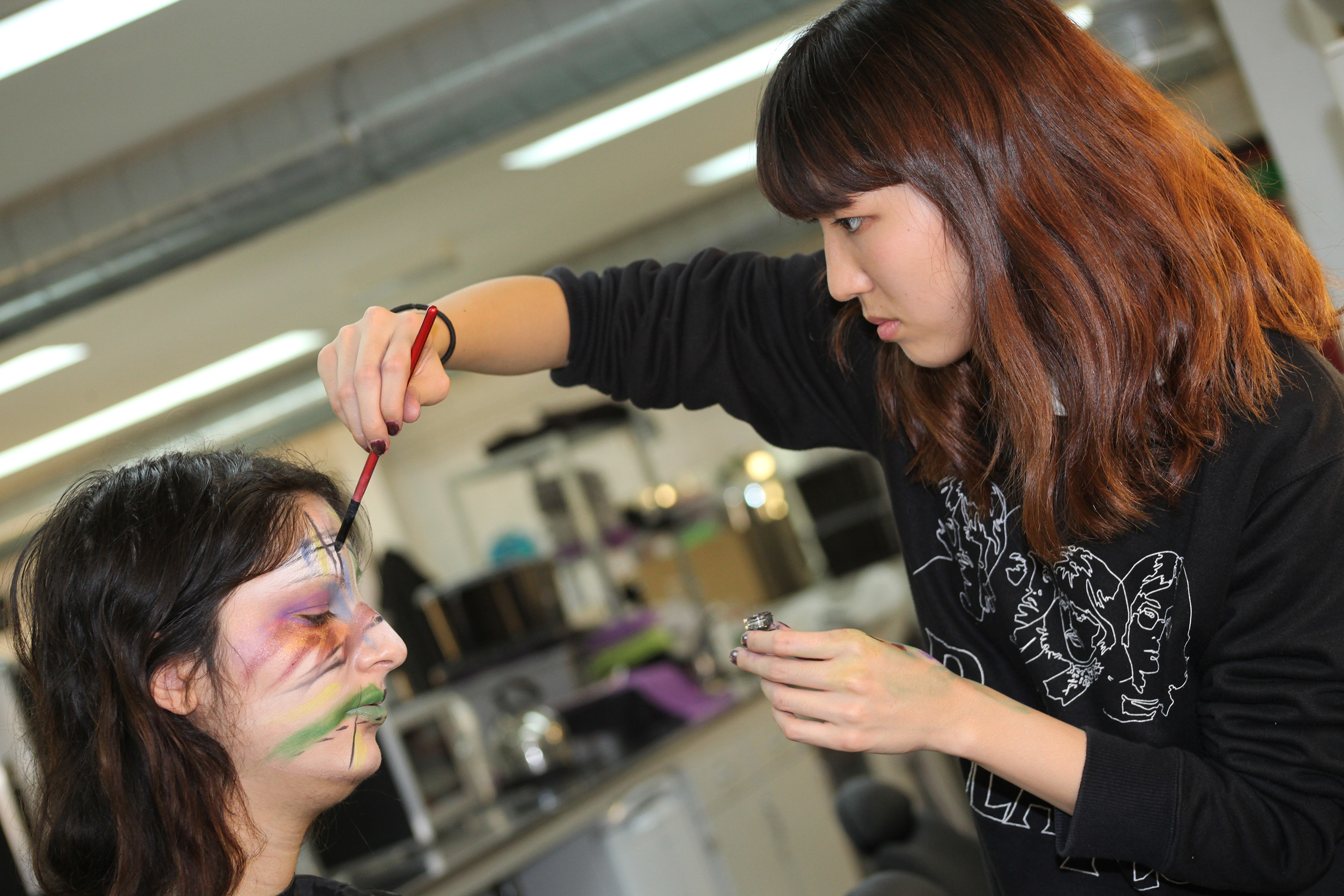 A student is painting and abstract design onto another student's face.