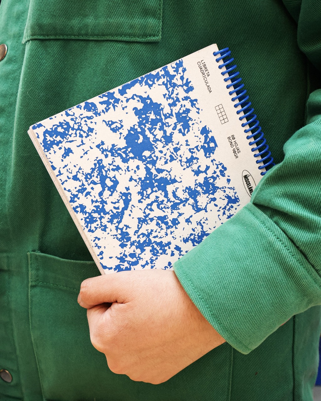 A person in a green coat holds a blue-and-white spiral notebook against their chest, with a pocket visible on the coat.