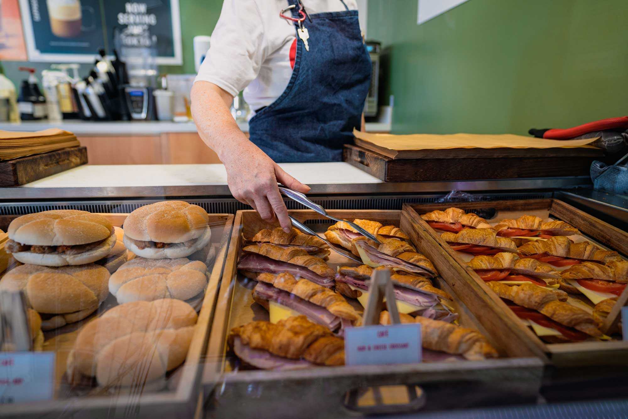 A person behind a counter prepares to pick up a ham and cheese croissant from a pile with a pair of tongs.