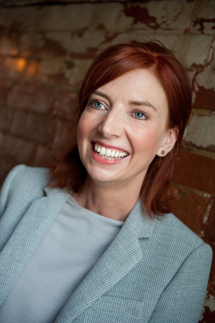 Woman with auburn hair and blue eyes smiling, wearing light blue jumper, sitting against exposed brick wall background.