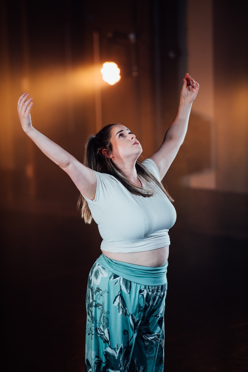 Woman with arms raised upwards wearing white crop top and teal patterned skirt, warm stage lighting creates orange glow in background.