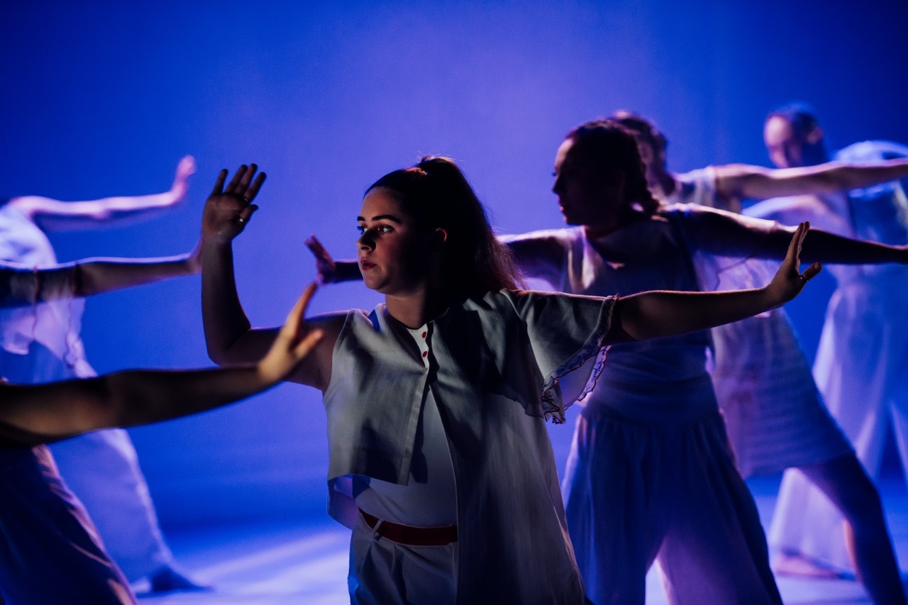 Woman in grey top with arms outstretched amongst dancers under blue and purple stage lighting.