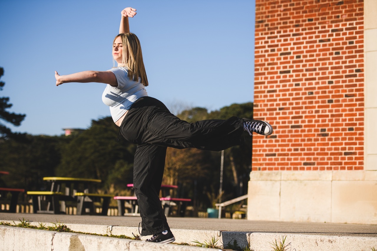 Woman in white top and dark trousers balancing on concrete ledge with one leg raised, brick building behind, blue sky visible.
