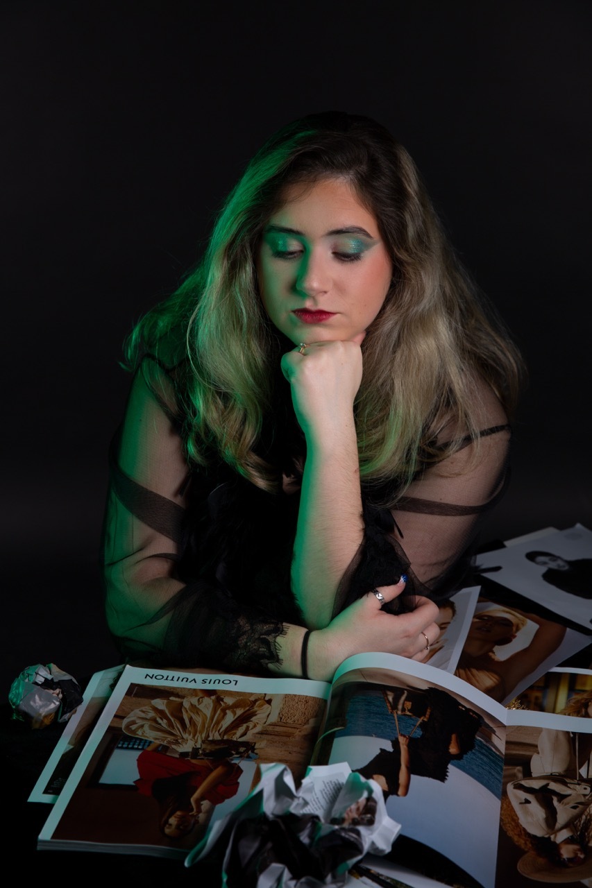Woman with blonde hair in black top, hand on chin, looking down at scattered photographs on table. Green lighting on left side.