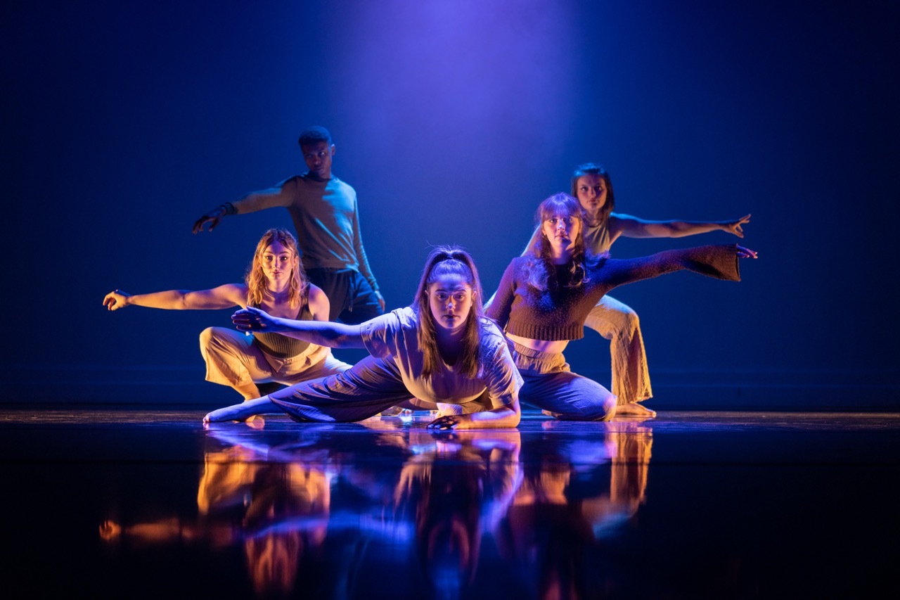 Four dancers on reflective floor in blue and purple stage lighting, with arms extended in synchronized poses.